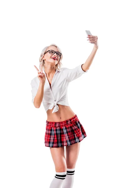 Соблазнительная блондинка в одежде школьницы показывает знак мира и делает селфи на смартфоне, изолированном на белом — стоковое фото