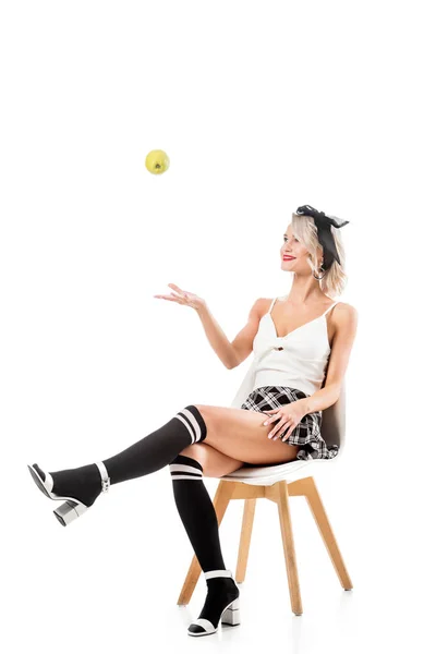 Young sexy woman in short plaid skirt and knee socks throwing apple while sitting on chair isolated on white — Stock Photo