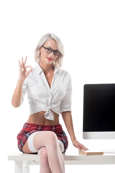 Blond seductive schoolgirl in short plaid skirt showing ok sign while sitting on table with blank computer screen isolated on white — Stock Photo