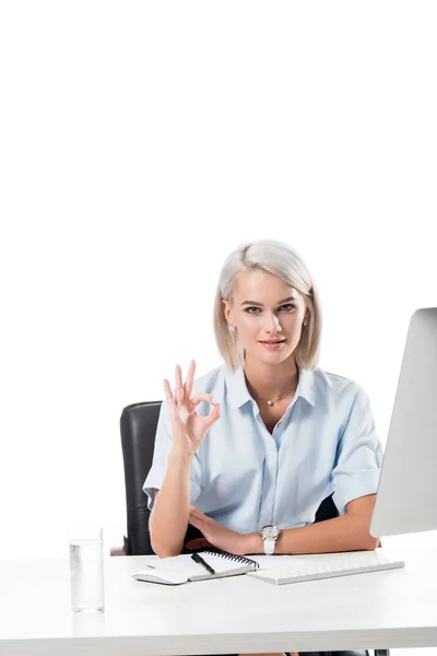 Porträt einer Geschäftsfrau mit Ok-Schild am Arbeitsplatz mit Glas Wasser, Notizbuch und Computerbildschirm auf weißem Hintergrund — Stockfoto