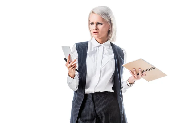 Portrait of confused businesswoman with notebook looking at smartphone in hand isolated on white — Stock Photo