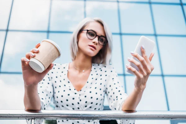 Focalizzata donna d'affari in occhiali con caffè per andare utilizzando smartphone sulla strada — Foto stock