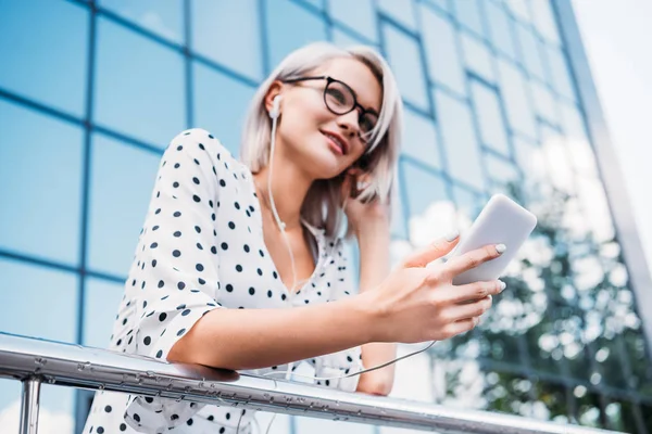 Niedrigwinkel-Ansicht der schönen Geschäftsfrau mit Smartphone in der Hand, die Musik in Kopfhörern auf der Straße hört — Stockfoto
