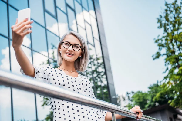 Mujer de negocios sonriente en gafas tomando selfie en teléfono inteligente en la mano en la calle - foto de stock