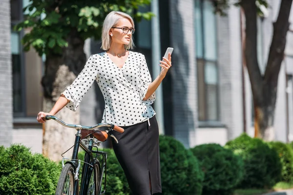 Beautiful businesswoman in eyeglasses using smartphone while leaning on bicycle on street — Stock Photo