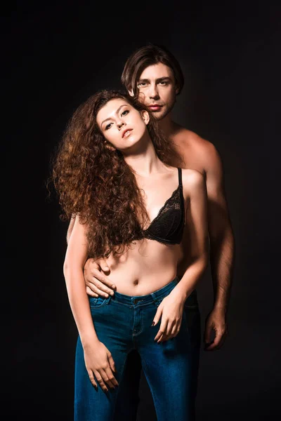 Portrait of man hugging girlfriend in black bra and jeans isolated on black — Stock Photo