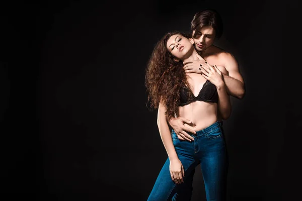 Man hugging seductive girlfriend in black bra and jeans isolated on black — Stock Photo