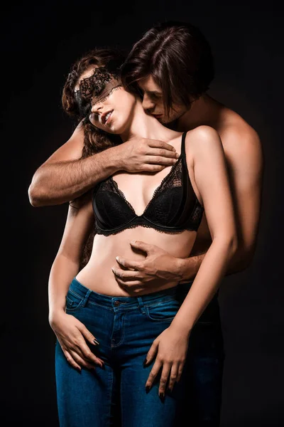 Retrato de hombre abrazando y besando sexy novia sin camisa con encaje negro en los ojos aislados en negro - foto de stock