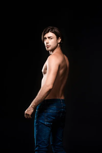 Back view of shirtless man in jeans looking at camera isolated on black — Stock Photo