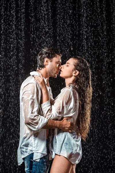 Vista lateral de pareja besándose bajo lluvia aislado en negro - foto de stock