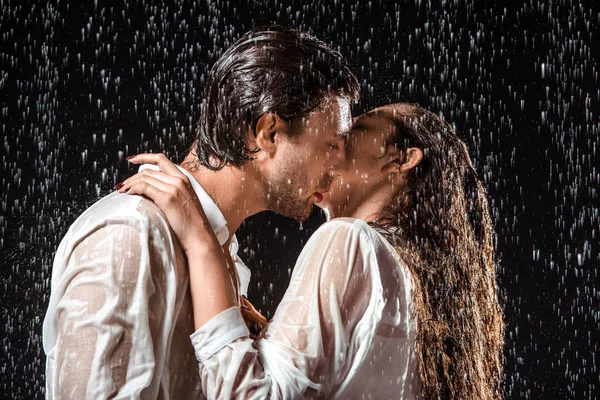 Vista lateral de pareja besándose bajo lluvia aislado en negro - foto de stock