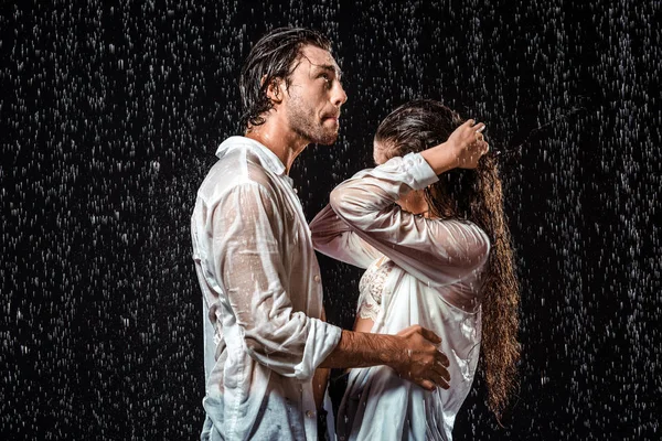 Vista lateral de pareja en camisas blancas de pie bajo la lluvia aisladas en negro - foto de stock