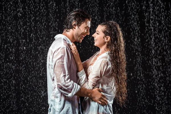 Vista lateral de la pareja sonriente abrazándose mientras está de pie bajo la lluvia aislado en negro - foto de stock