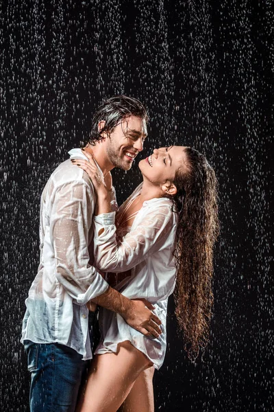 Vista lateral de casal sorridente abraçando enquanto em pé sob a chuva isolado em preto — Stock Photo