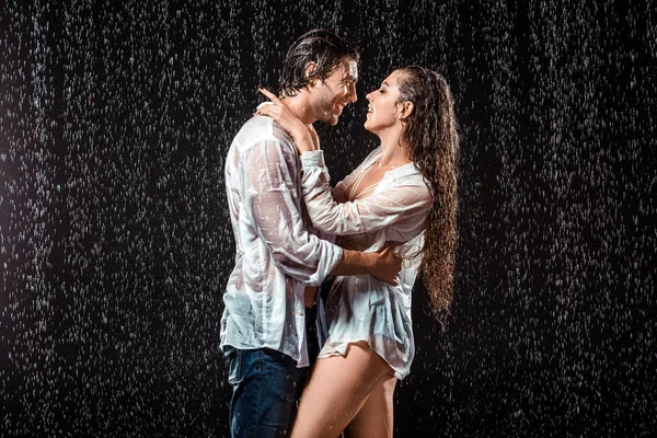 Vue latérale du couple souriant serrant dans ses bras sous la pluie isolé sur noir — Photo de stock