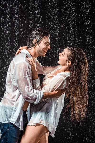 Happy wet couple in white shirts standing under rain isolated on black — Stock Photo