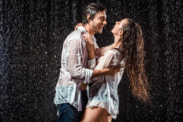 Feliz pareja mojada en camisas blancas de pie bajo la lluvia aislado en negro - foto de stock