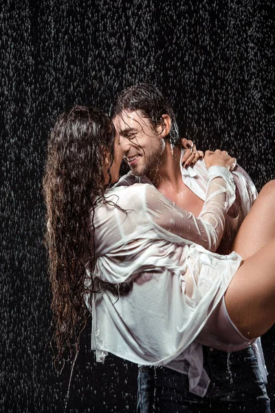 Smiling man holding girlfriend in white shirt while standing under rain isolated on black — Stock Photo