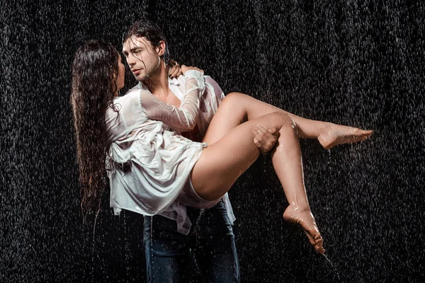Jovem homem segurando namorada em camisa branca, enquanto de pé sob a chuva isolado em preto — Fotografia de Stock