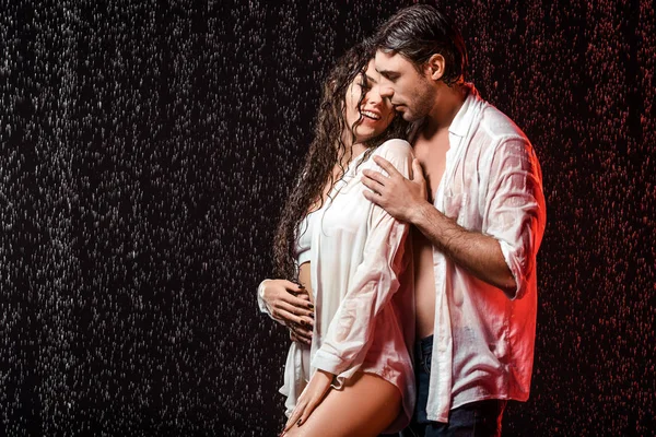 Portrait of sexy couple in white shirts standing under rain on black backdrop — Stock Photo