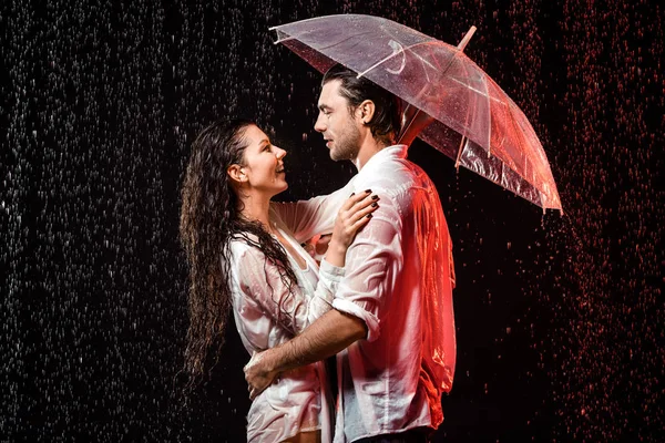 Vista laterale di coppia romantica in camicie bianche con ombrello in piedi sotto la pioggia su sfondo nero — Foto stock