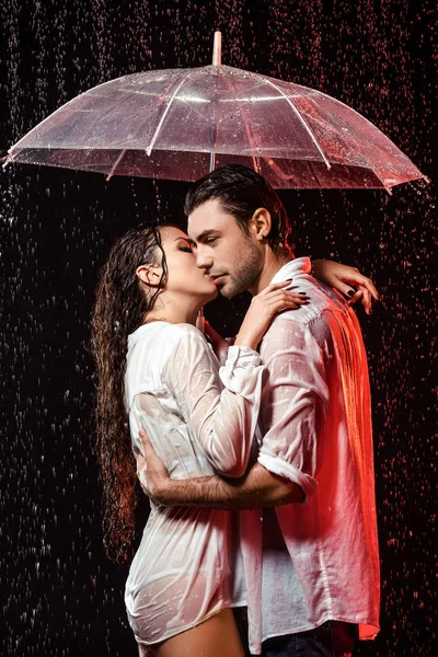 Вид сбоку романтической пары в белых рубашках с зонтиком, стоящей под дождем на черном фоне — стоковое фото
