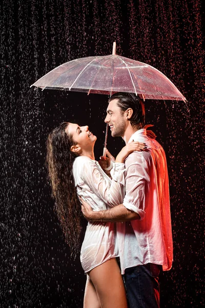 Vista lateral de pareja romántica en camisas blancas con paraguas de pie bajo la lluvia sobre fondo negro - foto de stock
