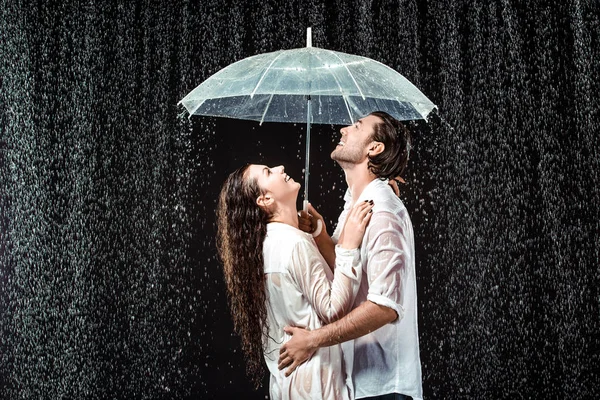Vista lateral de feliz pareja en camisas blancas de pie bajo paraguas bajo gotas de lluvia aisladas en negro - foto de stock