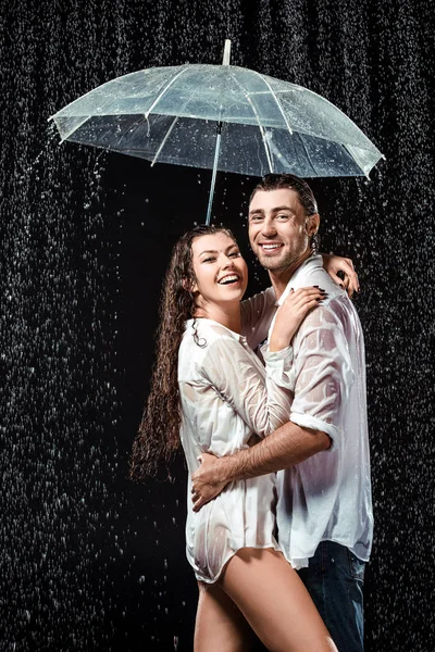 Vista lateral de feliz pareja en camisas blancas de pie bajo paraguas bajo gotas de lluvia aisladas en negro - foto de stock