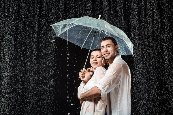 Vista lateral de pareja romántica en camisas blancas de pie bajo paraguas bajo gotas de lluvia aisladas en negro - foto de stock