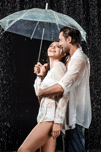 Vista lateral de pareja romántica en camisas blancas de pie bajo paraguas bajo gotas de lluvia aisladas en negro - foto de stock