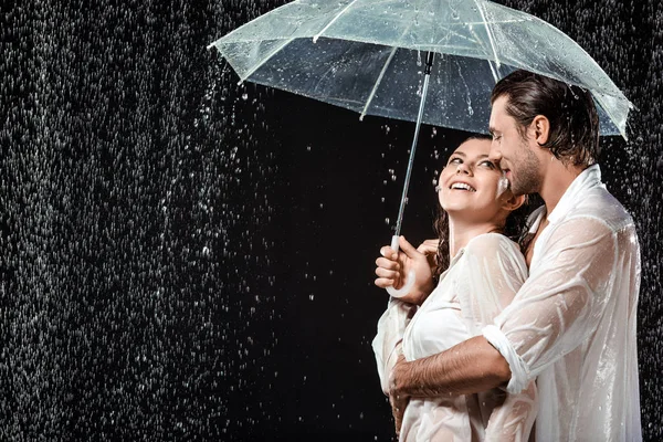 Vista lateral de pareja romántica en camisas blancas de pie bajo paraguas bajo gotas de lluvia aisladas en negro - foto de stock