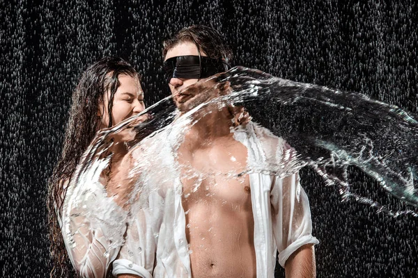 Man with black ribbon on eyes and girlfriend while swilled with water isolated on black — Stock Photo