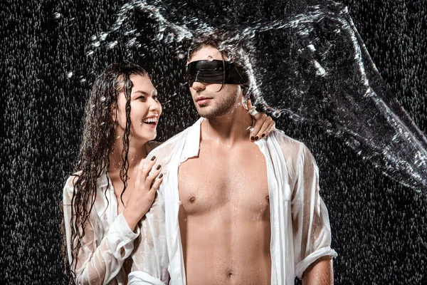 Ritratto di uomo con nastro nero sugli occhi e fidanzata sorridente mentre scorreva con acqua isolata sul nero — Foto stock