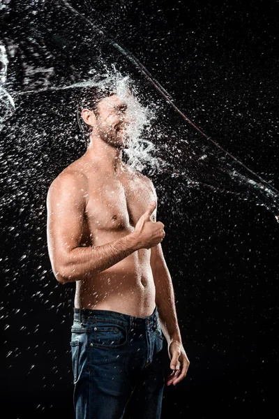 Sonriente joven mostrando el pulgar hacia arriba mientras hinchado con agua aislada en negro - foto de stock