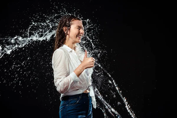 Vue latérale de la femme souriante en chemise blanche montrant pouce vers le haut tandis que swilled avec de l'eau isolée sur noir — Photo de stock