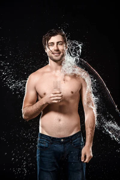 Retrato del hombre sin camisa sonriente con vaso de agua en la mano y salpicaduras de agua aisladas en negro - foto de stock