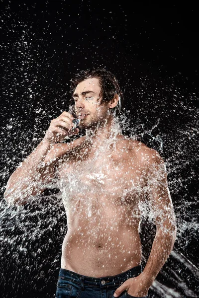 Retrato del hombre sin camisa bebiendo agua de vidrio mientras se hincha con agua salpicada aislada en negro - foto de stock