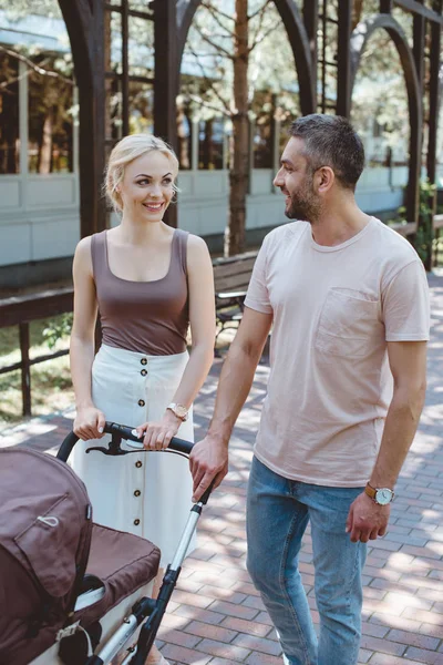 Smiling parents walking with baby carriage in park and looking at each other — Stock Photo