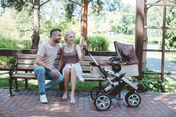 Pai e mãe sentados no banco perto da carruagem do bebê no parque — Fotografia de Stock