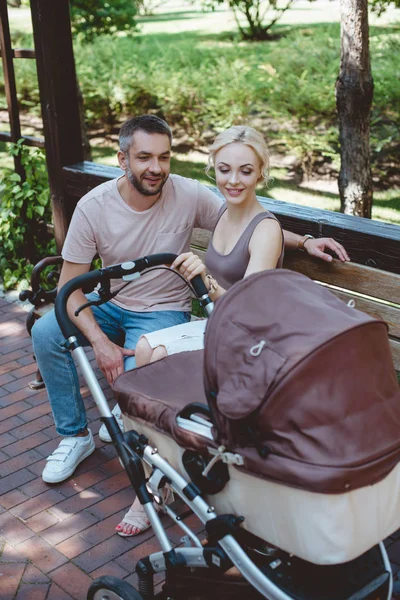 Pais sentados no banco perto da carruagem do bebê e olhando para a criança no parque — Fotografia de Stock