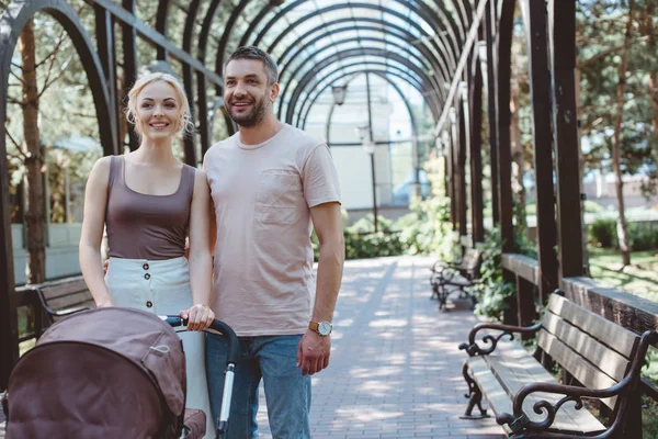 Smiling parents standing with baby carriage in park and looking away — Stock Photo