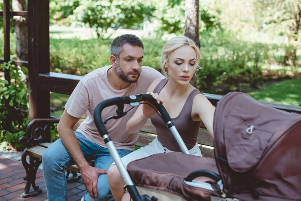 Pais sentados no banco e olhando para a carruagem do bebê no parque — Fotografia de Stock