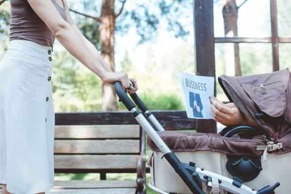 Immagine ritagliata della donna che tiene la carrozza del bambino nel parco, qualcuno che tiene il giornale di affari nella carrozza del bambino — Foto stock