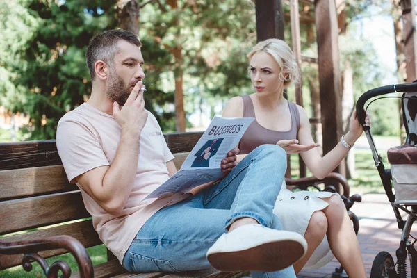 Husband smoking cigarette and reading newspaper near baby carriage in park, angry wife looking at him — Stock Photo