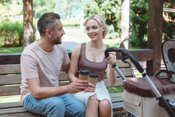 Eltern sitzen auf Bank neben Kinderwagen im Park und halten Kaffee in Pappbechern — Stockfoto