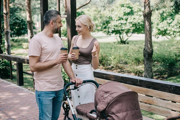 Pais alegres segurando café para ir perto da carruagem do bebê no parque — Fotografia de Stock