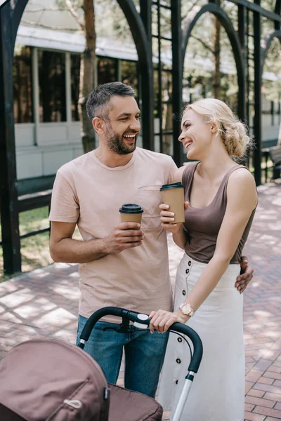 Pais rindo segurando café para ir perto de carruagem do bebê no parque — Fotografia de Stock