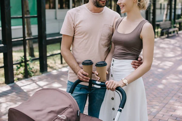 Cropped image of parents holding coffee to go near baby carriage in park — Stock Photo