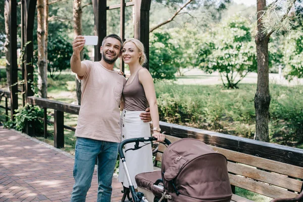 Madre y padre tomando selfie con teléfono inteligente cerca de carro de bebé en el parque - foto de stock
